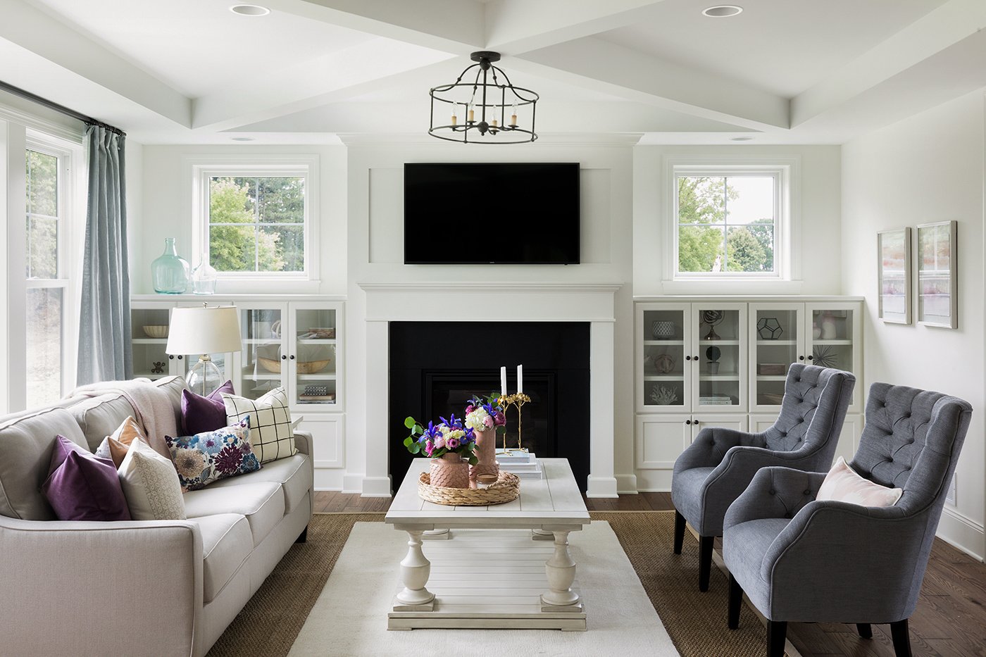A cottage-style living room with a fireplace flanked by picture windows, and a couch under a row of single-hung windows.